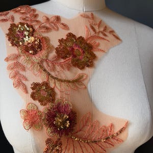 Può includere: Un'applicazione in tessuto a rete color pesca con motivi floreali ricamati in tonalità corallo, rosa e oro. Il design presenta un intricato lavoro di fili e paillettes dorate metallizzate disposte in motivi di fiori e foglie sulla base.