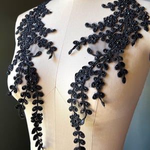 May include: Black lace applique with a floral vine design. The applique features small, dark beads and is displayed on a mannequin. The delicate design is suitable for embellishing garments or accessories.