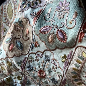 Puede incluir: Primer plano de tela bordada con motivos florales y cachemir. La paleta de colores incluye crema, azul claro, melocotón y verdes suaves. El bordado en relieve crea un efecto texturizado. La tela sugiere un material de alta calidad, posiblemente seda.