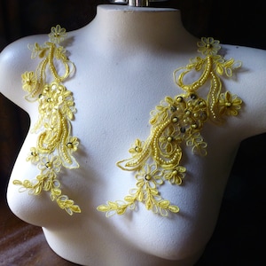 May include: Two yellow lace appliques with floral and beaded designs. The appliques feature delicate leaves, small flowers, and beaded accents. They are displayed on a white mannequin torso, suggesting they are for clothing embellishment. The background is dark.
