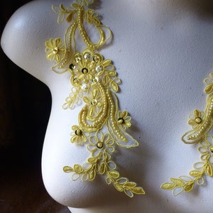 May include: Close-up of a white mannequin torso adorned with two yellow lace appliques. The appliques feature floral and leaf designs, embellished with sequins and small beads. The lace is a bright, sunny yellow.