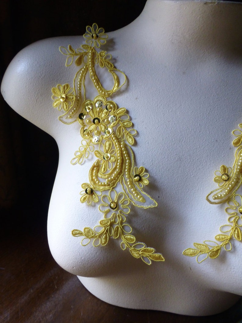 May include: Close-up of a white mannequin torso adorned with two yellow lace appliques. The appliques feature floral and leaf designs, embellished with sequins and small beads. The lace is a bright, sunny yellow.