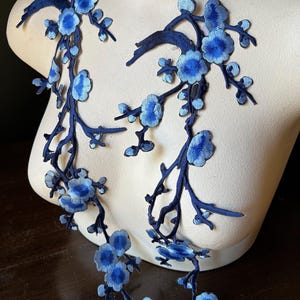 Pode incluir: Apliques florais bordados em tons de azul. Os apliques apresentam costuras detalhadas de flores e galhos, criando um design delicado e ornamental. Os apliques são exibidos em um manequim, mostrando seu uso potencial em projetos de moda ou artesanato.