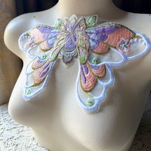 Puede incluir: Una delicada aplicación en forma de mariposa con un diseño floral en tonos rosa, morado, verde y dorado. La aplicación está hecha de tela transparente y tiene un fondo blanco.