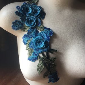 Puede incluir: Una aplicación floral azul y verde con tres rosas grandes y flores más pequeñas. La aplicación está hecha de tela y es perfecta para agregar un toque de elegancia a cualquier proyecto.