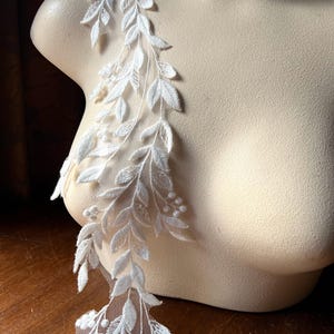 May include: White floral lace applique featuring a delicate leaf and berry design. The applique is attached to a sheer mesh backing, creating a flowing, elegant look. Perfect for embellishing bridal wear or other craft projects.