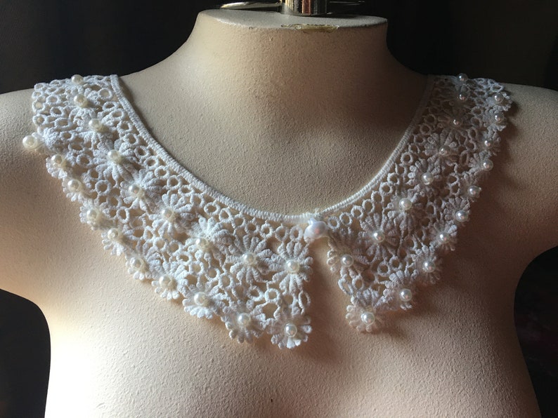 Puede incluir: Collar de encaje blanco de ganchillo con adornos de perlas. El collar tiene un borde festoneado y un centro puntiagudo.