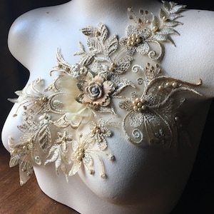 May include: A gold and ivory floral lace applique with pearl beads. The applique is on a white mannequin bust.