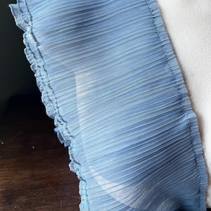 May include: Close-up of a light blue, pleated fabric with a ruffled edge. The fabric appears to be sheer and delicate, with a subtle texture. The image highlights the intricate details of the material.