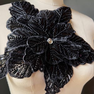 May include: A black floral applique with intricate sequin and bead detailing. The applique features layered petals and a central rhinestone embellishment. This decorative piece is likely for embellishing garments or accessories.