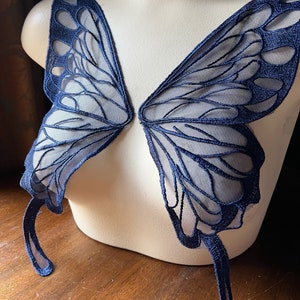 Op de afbeelding: Een paar donkerblauwe vlinder vleugels gemaakt van transparante stof met geborduurde details. De vleugels zijn bevestigd aan een wit mannequin.