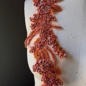 May include: An ornate, orange and red beaded applique, featuring a floral design with intricate detailing. The applique is composed of various sizes of sparkling beads and crystals, creating a dimensional and textured effect. It is attached to a sheer, flesh-toned fabric.