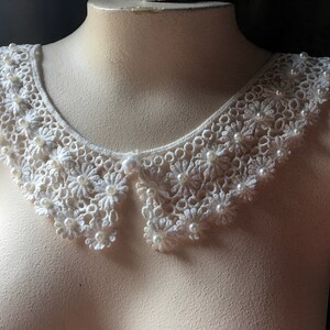 Puede incluir: Un cuello de encaje blanco con adornos de perlas. El cuello es delicado y femenino, y ser&iacute;a perfecto para agregar un toque de elegancia a cualquier atuendo.
