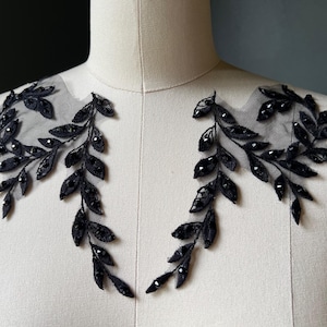 Puede incluir: Dos apliques de hojas negras con cuentas sobre tela transparente, exhibidos en un maniquí. Los apliques presentan diseños de hojas detallados con cuentas oscuras, ideales para adornar prendas o accesorios. El diseño es elegante.