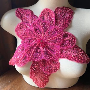Pode incluir: Uma vibrante aplicação floral rosa, ricamente adornada com lantejoulas e contas. A aplicação é exibida em um manequim, mostrando seu design e textura. A flor tem múltiplas pétalas e um centro detalhado.