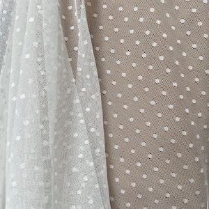 May include: Close-up of white sheer fabric with a polka dot pattern. The fabric is draped, showing the texture and transparency of the material. The dots are evenly spaced and create a delicate, airy appearance.