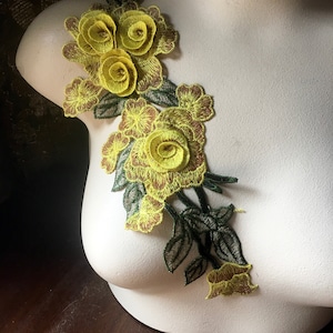 Puede incluir: Aplique de bordado floral amarillo con hojas verdes. El aplique presenta múltiples capas de flores de tela amarilla con costuras marrones.