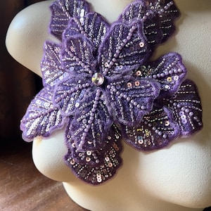 May include: A large, purple floral applique with intricate detailing. The petals are embellished with sequins and beads, radiating from a central crystal. The applique is displayed on a mannequin, showcasing its design and texture.