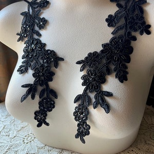 May include: Two black floral lace appliques with beaded accents are displayed on a white mannequin. The appliques feature detailed floral and leaf patterns, ideal for embellishing garments or crafts. The lace is intricately designed.