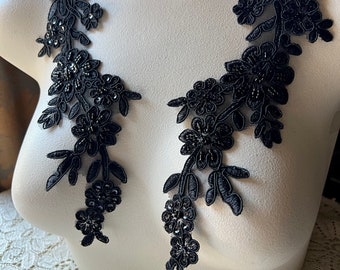 May include: Two black floral lace appliques with beaded accents are displayed on a white mannequin. The appliques feature detailed floral and leaf patterns, perfect for embellishing garments or crafts. The lace is intricately designed.