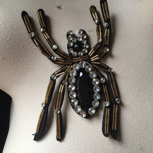 May include: A black and gold spider-shaped applique with rhinestones. The spider has a black velvet body and gold bead legs.