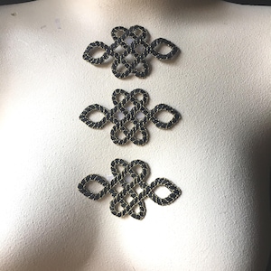 May include: Three decorative black and gold knot closures are displayed on a mannequin. The closures are made of interwoven black material with gold trim, and are arranged vertically. The background is a neutral off-white.
