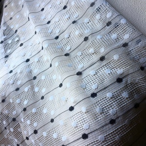 May include: Close-up of a white lace fabric with a grid pattern. Black and white polka dots are arranged along diagonal lines. The fabric has a delicate, open weave, creating a textured surface. The overall effect is elegant and airy.