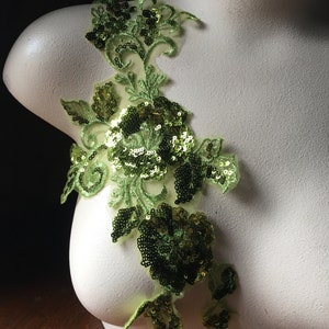 Può includere: Applique floreale verde con paillettes. L'applique presenta un grande fiore con fiori e foglie più piccoli. L'applique è realizzata in un tessuto trasparente con paillettes cucite.