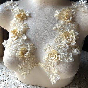 May include: Ivory lace applique with 3D floral and leaf details. The applique features multiple clusters of delicate, cream-colored roses and leaves, perfect for embellishing garments or crafts. The lace is attached to a mannequin.