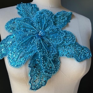 May include: A large, turquoise floral applique, heavily embellished with sequins and beads. The flower is displayed on a mannequin, showcasing its intricate design and potential use in fashion or craft projects. The petals are layered and detailed.