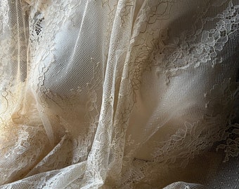 May include: Ivory-colored lace fabric draped over a mannequin. The delicate lace features intricate floral patterns and a sheer, open weave. The fabric is softly lit, highlighting the texture and design. Ideal for bridal or vintage-inspired projects.