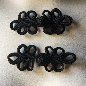 May include: Two black Chinese knot buttons, also known as frog closures, are shown on a white surface. The knots are intricately woven and have a decorative, floral design.