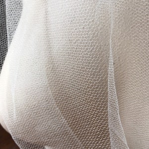 May include: Close-up of white tulle fabric with a hexagonal mesh pattern. The fabric is draped, creating soft curves and shadows. The texture is fine and delicate, suggesting a lightweight material.