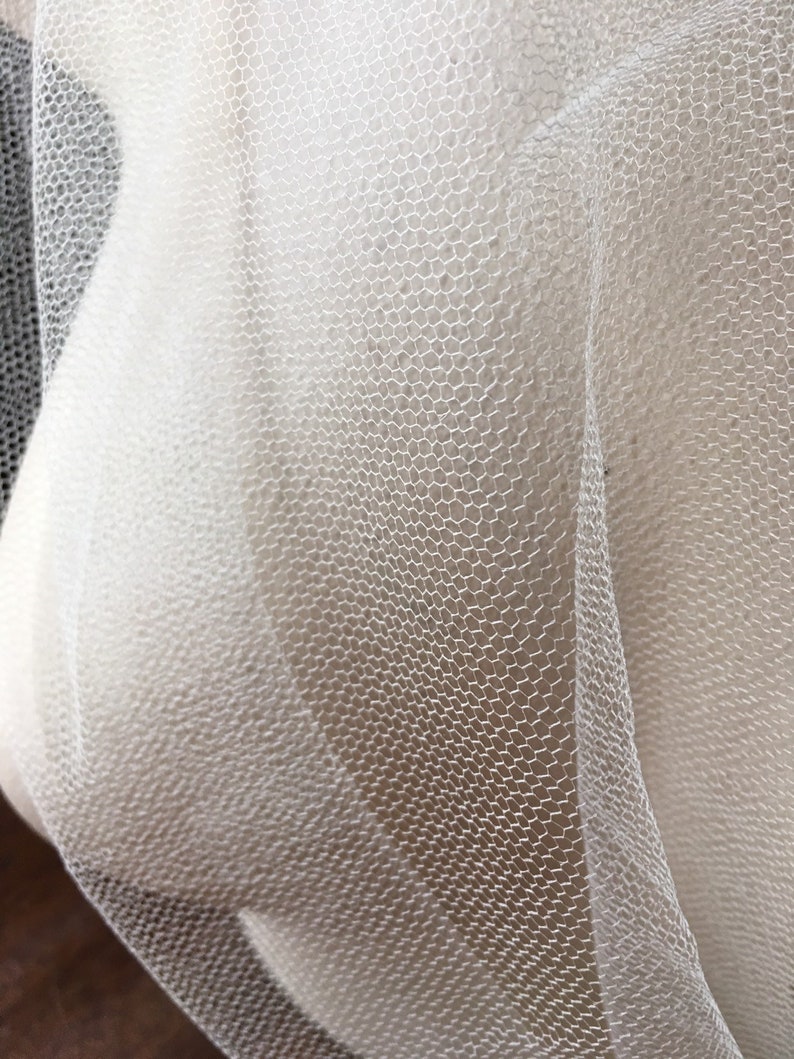 May include: Close-up of white tulle fabric with a hexagonal mesh pattern. The fabric is draped, creating soft curves and shadows. The texture is fine and delicate, suggesting a lightweight material.