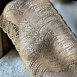 May include: Close-up of a gold-colored lace trim with a scalloped edge. The lace features a repeating pattern of stylized leaves and floral elements. The trim is draped over a mannequin, showcasing its delicate design and texture.