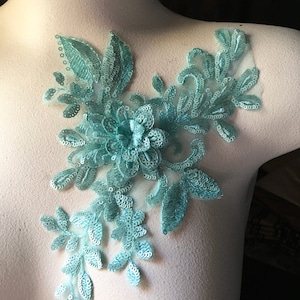 May include: A light blue floral applique with sequins and lace. The applique features a large flower in the center surrounded by smaller leaves.