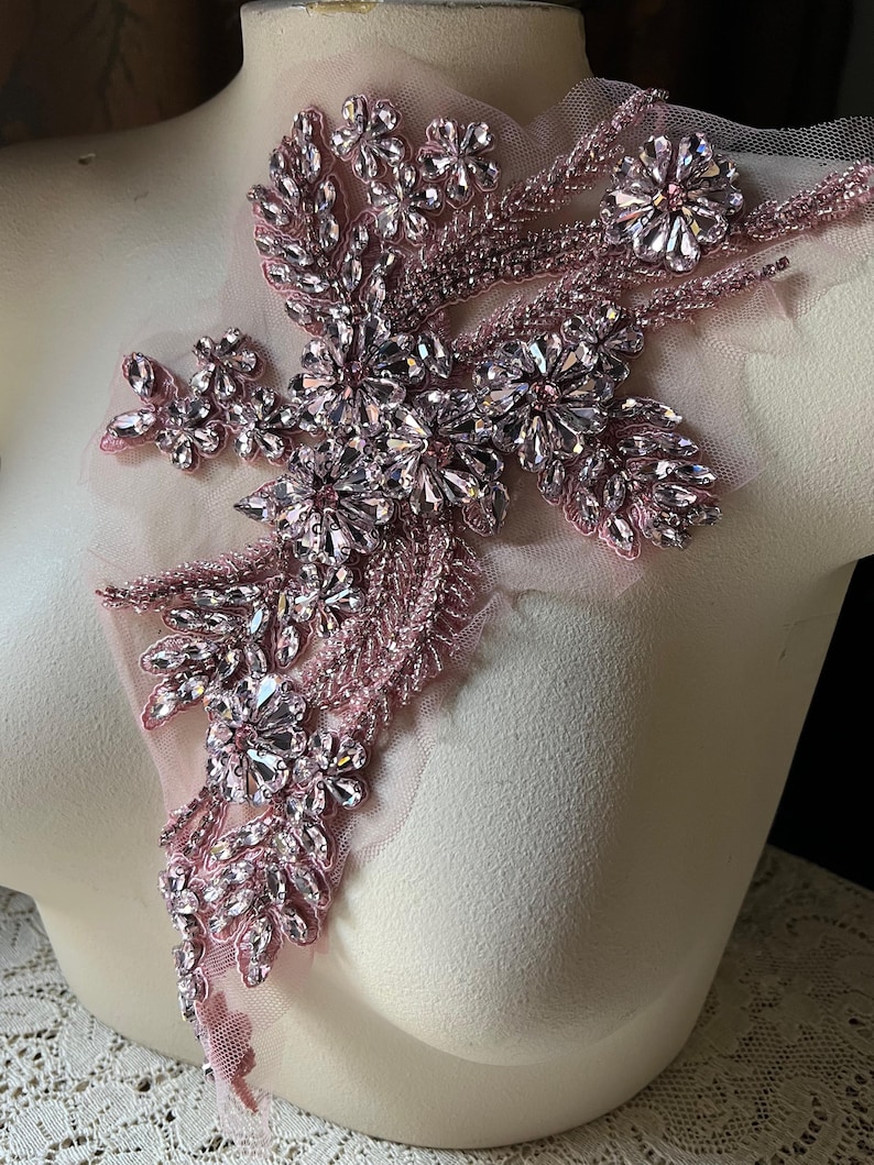 Puede incluir: Una aplicaci&oacute;n de cuentas y diamantes de imitaci&oacute;n de color rosa con un dise&ntilde;o floral. La aplicaci&oacute;n est&aacute; sobre un fondo de tela transparente.