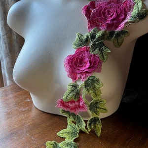 Op de afbeelding: Een roze en groene bloemenapplicatie met drie roze rozen en groene bladeren. De applicatie is gemaakt van stof en is bedoeld om op kleding of andere stoffen items te worden genaaid.