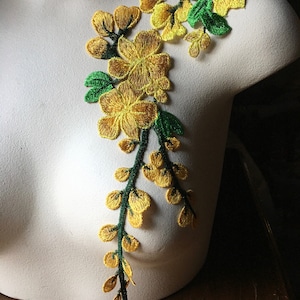 May include: Embroidered floral applique featuring yellow flowers and green leaves on a dark green stem. The applique is displayed on a white mannequin, showcasing the intricate details of the design. This decorative element is suitable for embellishing garments or crafts.