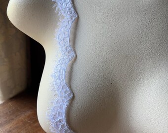 May include: White lace trim with a scalloped edge, displayed on a mannequin. The delicate lace has a detailed, openwork design and a fringed edge. The trim is likely used for sewing, crafting, or adding a decorative touch to garments or home decor.