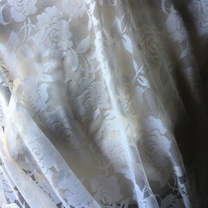 May include: Off-white lace fabric with a floral rose pattern. The delicate material is draped, creating soft folds and shadows. The lace is semi-transparent, revealing a lighter background.