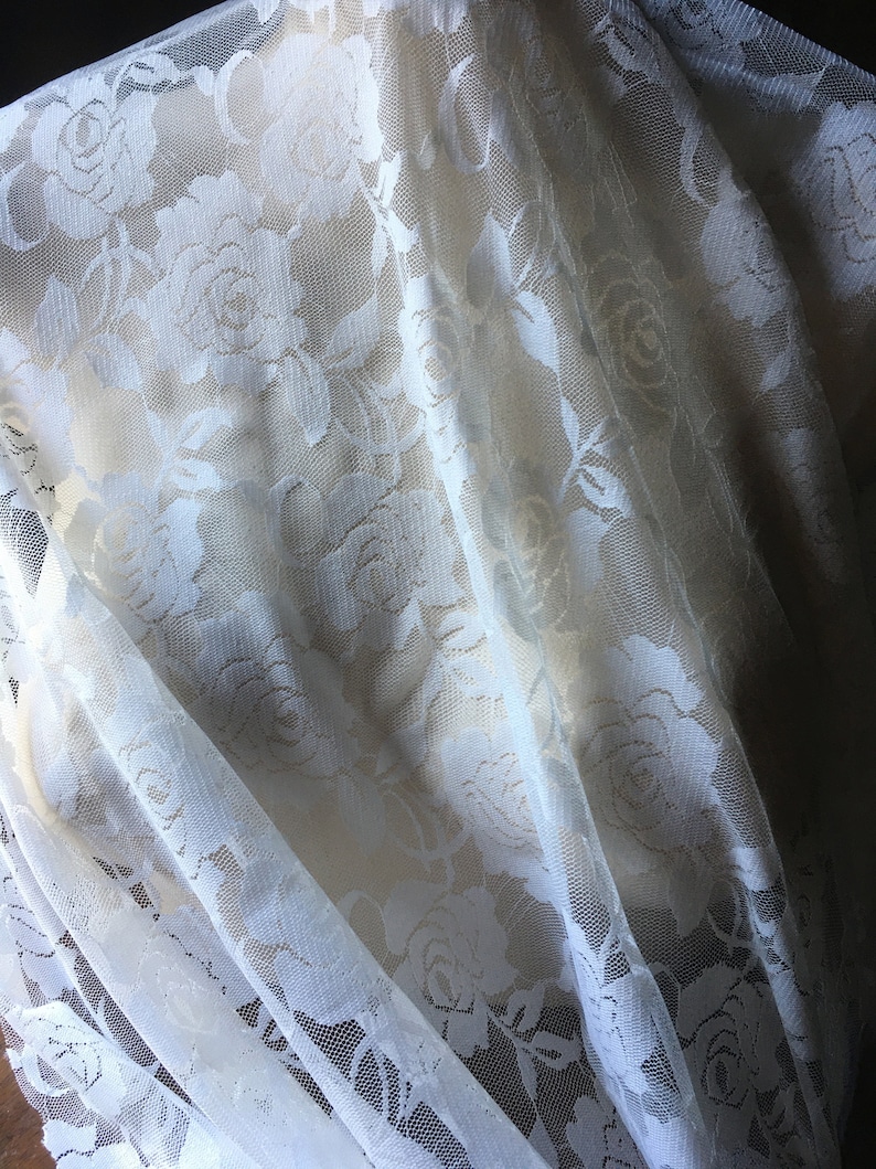 May include: Off-white lace fabric with a floral rose pattern. The delicate material is draped, creating soft folds and shadows. The lace is semi-transparent, revealing a lighter background.