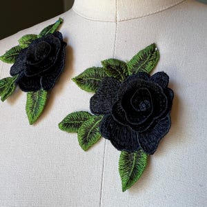Puede incluir: Dos apliques de rosas negras con hojas verdes. Las rosas bordadas tienen pétalos detallados y están rodeadas de hojas verdes con costuras intrincadas. Ideales para añadir un toque decorativo a la ropa o accesorios.