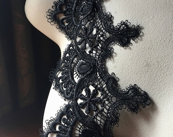 May include: Black lace trim with an intricate floral design. The trim is displayed on a mannequin, showcasing the delicate openwork and detailed patterns. The lace is a deep black color.