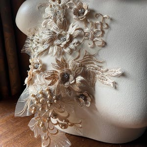 May include: A delicate gold lace applique with pearl embellishments and floral designs. The applique is perfect for adding a touch of elegance to wedding dresses, bridal accessories, or other craft projects.