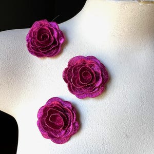 Puede incluir: Tres apliques de rosas de tela fucsia vibrante. Las rosas están bordadas con un hilo metálico, creando un efecto brillante. Los apliques están dispuestos sobre una superficie blanca, mostrando sus intrincados detalles y su rico color.