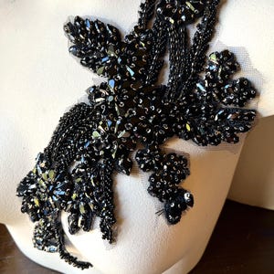 Könnte beinhalten: Schwarze Perlenapplikation mit floralem Design. Die Applikation weist eine Vielzahl schwarzer Perlen und Pailletten auf, die einen strukturierten und dimensionalen Effekt erzeugen. Das Design ist auf einem transparenten Mesh-Träger befestigt.