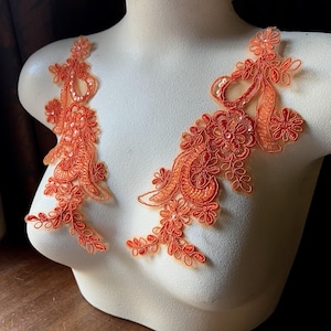 May include: Two orange floral lace appliques with sequin accents. The appliques are displayed on a mannequin, showcasing their intricate design and potential placement on garments. The lace features delicate floral patterns and a vibrant orange hue.