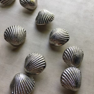 Puede incluir: Una colección de objetos decorativos plateados con forma de concha. Los objetos tienen una textura estriada y están dispuestos sobre una tela de color claro. Las conchas probablemente se utilizan para la decoración del hogar o proyectos de artesanía.