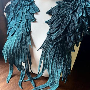 May include: Two teal green fabric wings with a layered feather design. The wings are attached to a white mannequin.